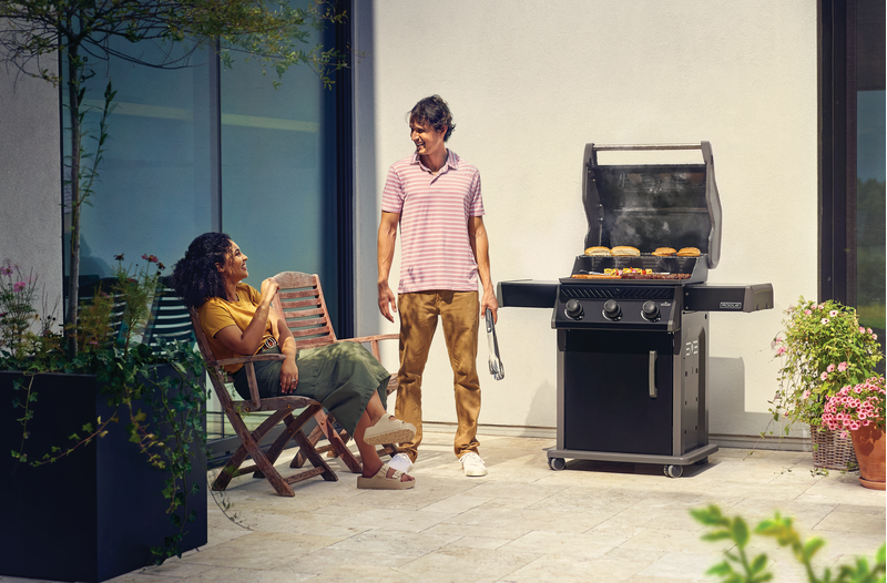 Napoleon Rogue 425 gas grill in use on backyard patio with food cooking, foldable side shelves extended, and sleek black porcelain enamel finish visible